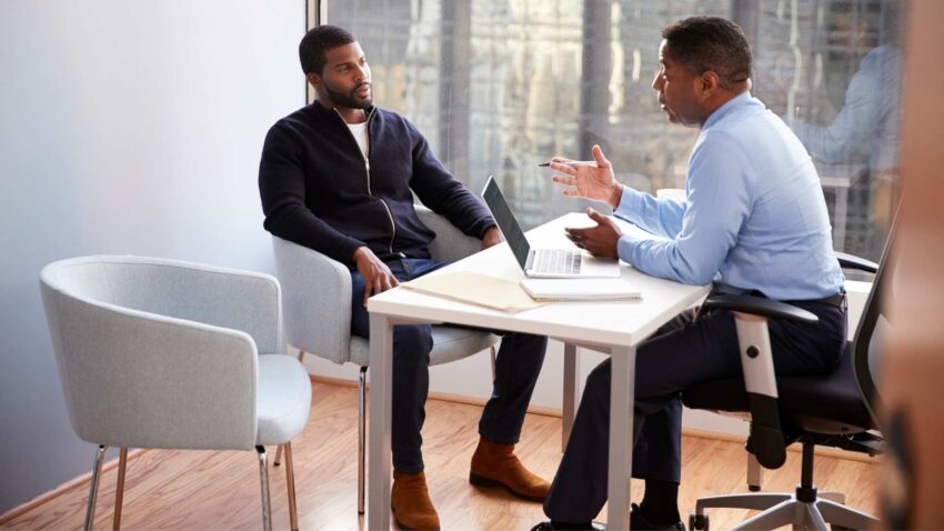 Deux hommes assis autour d'une table, l'un consultant un ordinateur tout en discutant avec l'autre