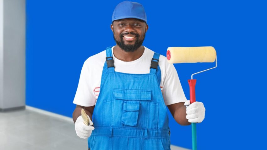 Peintre en tenue de travail devant un mur bleu, tenant pinceau et rouleau