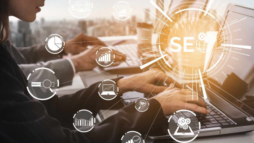 Inscription SEO avec ces domaines en avant et en arrière-plan; deux personnes travaillant sur ordinateur